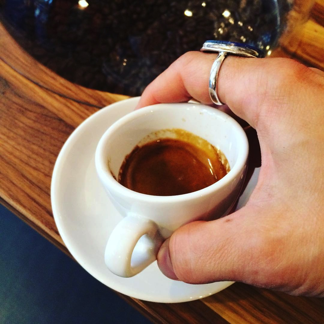 A hand holding a white espresso cup containing Valmandin's Baba Budan espresso freshly brewed and with a thick crema.
